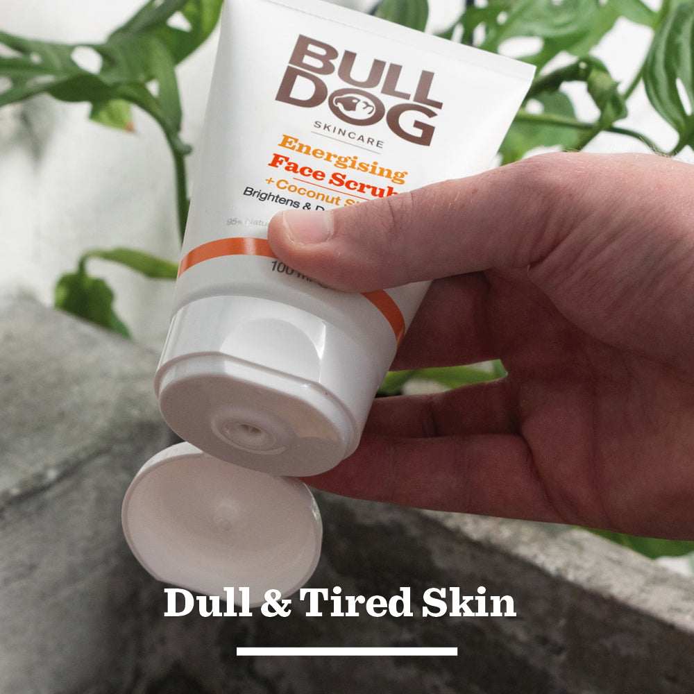 A hand squeezes a tube of Energising Face Scrub for men, made with natural origin ingredients, against green plants and concrete. Text overlay: “Dull & Tired Skin.”.
