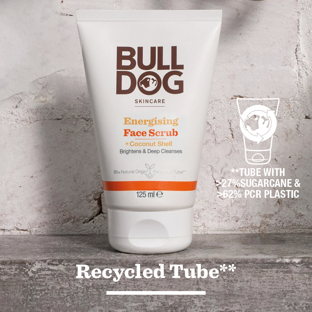 A tube of Energising Face Scrub stands upright on a gray surface. Text notes it contains 27% sugarcane and 62% post-consumer recycled plastic, and is made with natural origin ingredients. “Recycled Tube**” appears below.