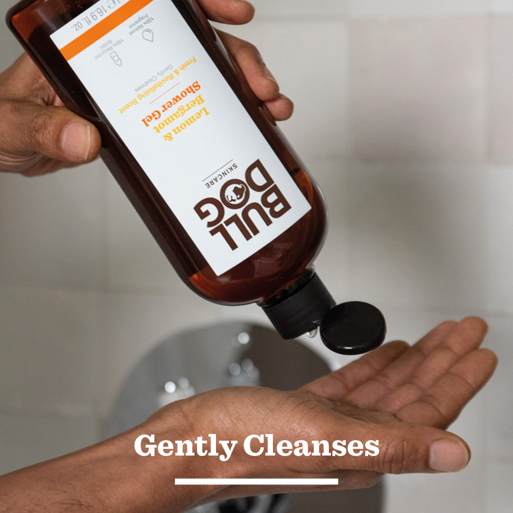 A person sprays Energising Spray Deodorant Bundle from a Bull Dog Skincare bottle under their arm, standing by a sink. Text at the bottom reads Gently Refreshes.