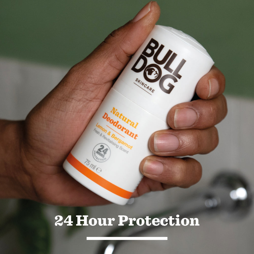 A hand holds a white stick of Lemon & Bergamot Natural Deodorant. “24 Hour Protection” is shown underneath, highlighting this men’s natural deodorant. The background features a blurred green and white blend.