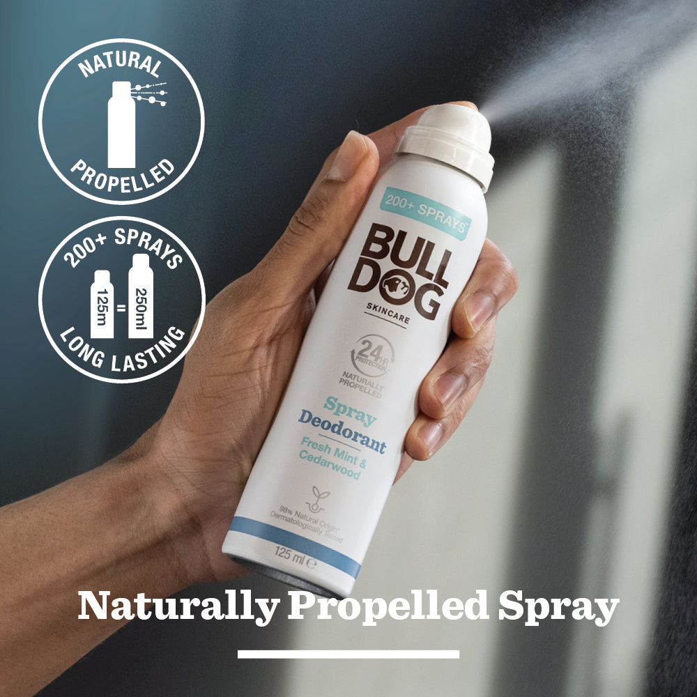 A hand sprays Bulldog’s Fresh Mint & Cedarwood Spray Deodorant. The label highlights 24-hour odor protection, 200+ sprays, and fresh scent. Circular icons note “Natural Propelled,” “Long Lasting,” and natural origin ingredients.