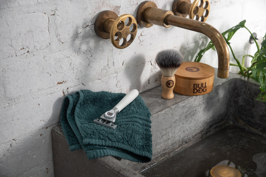 A brass faucet and handles are above a concrete sink where a green towel, Sensitive Steel Blades razor, shaving brush, and wooden BULLDOG soap bowl rest. A green plant is partially visible on the right.