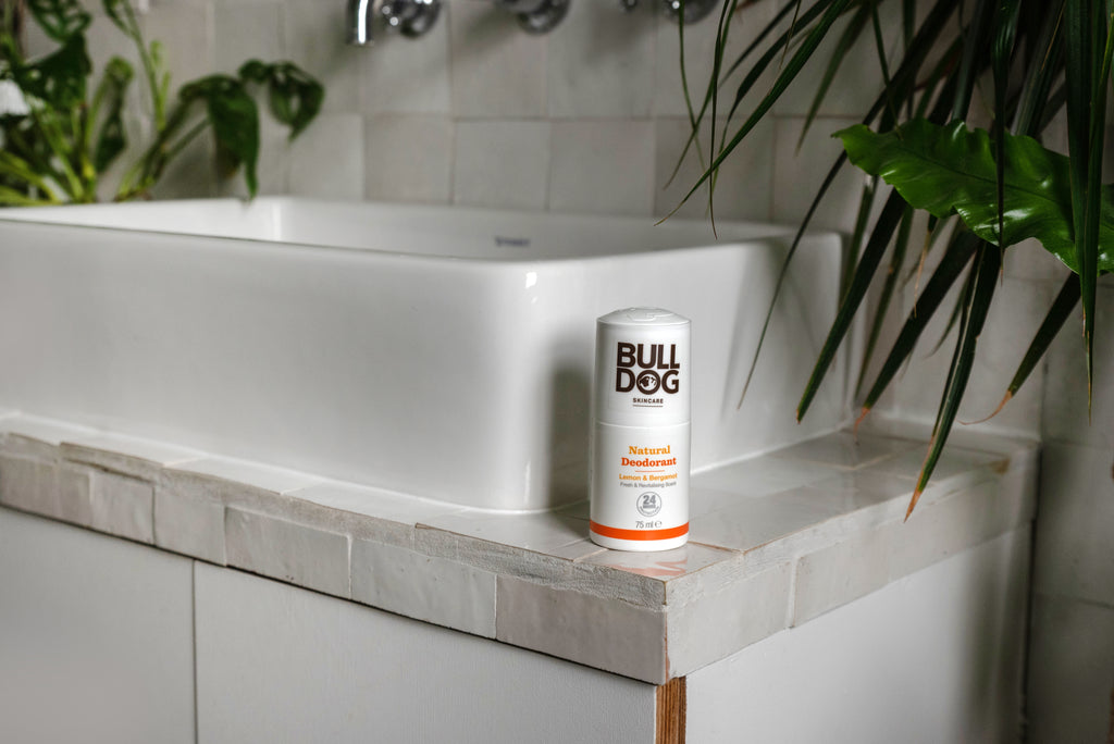 A stick of Lemon & Bergamot Natural Deodorant, offering 24-hour odour protection, sits on the tiled edge of a white bathroom sink, surrounded by green plants.
