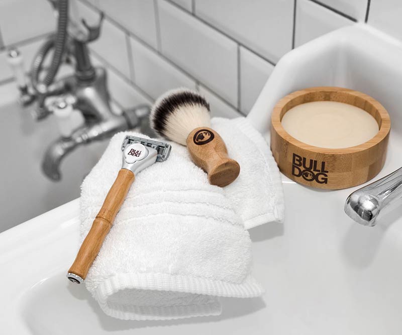 The Premium Shave Collection Bundle rests on a folded white towel beside a white sink, tiled walls, and a metal faucet.