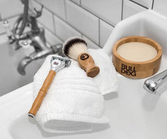 The Premium Shave Collection Bundle rests on a folded white towel beside a white sink, tiled walls, and a metal faucet.