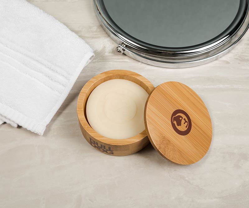 A round bar of soap from our Premium Shave Collection Bundle sits in a bamboo container with the lid off, placed on a marble countertop next to a folded white towel and a round mirror.
